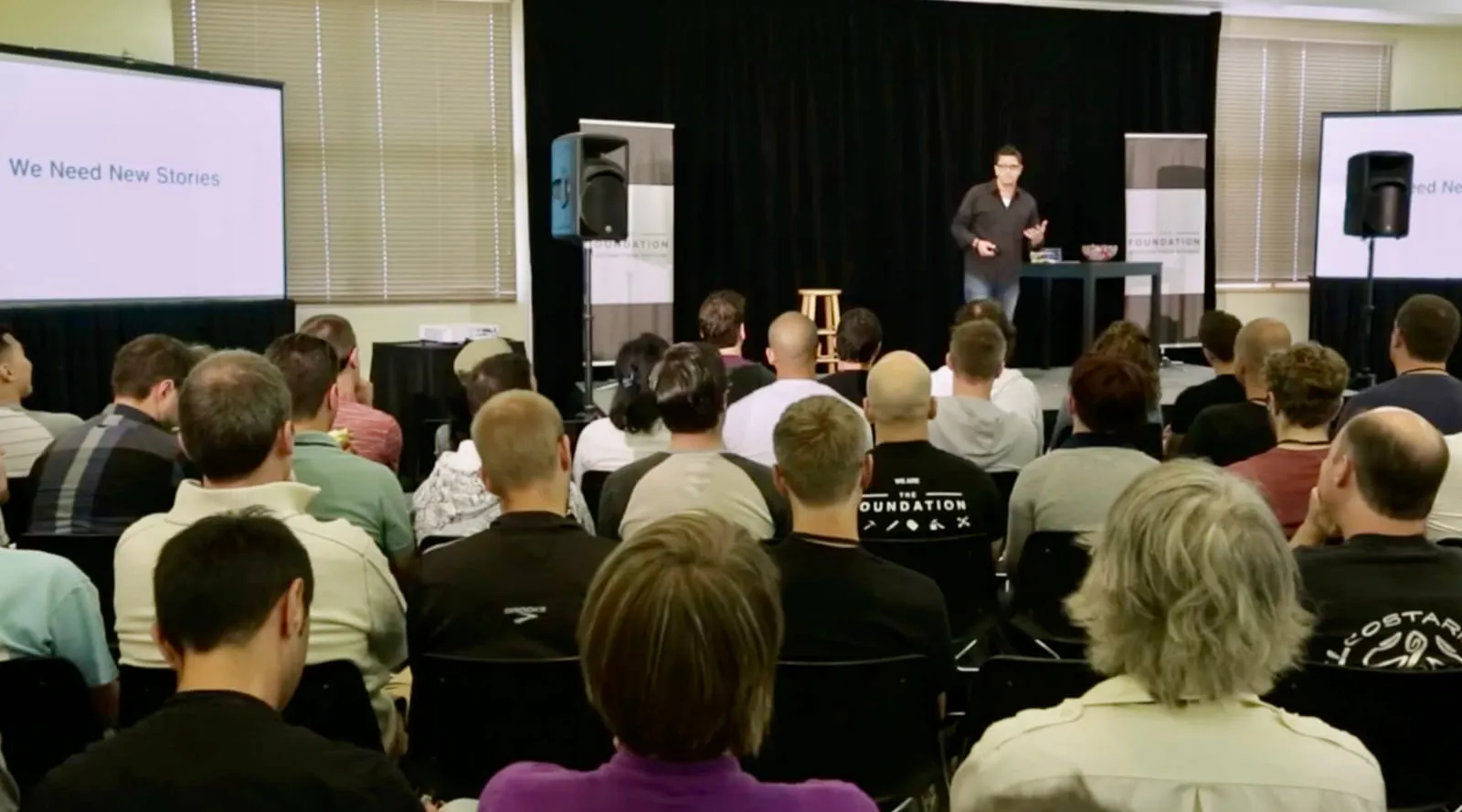 Rob Scott presenting to a large audience, with "We Need New Stories" on the screen behind him